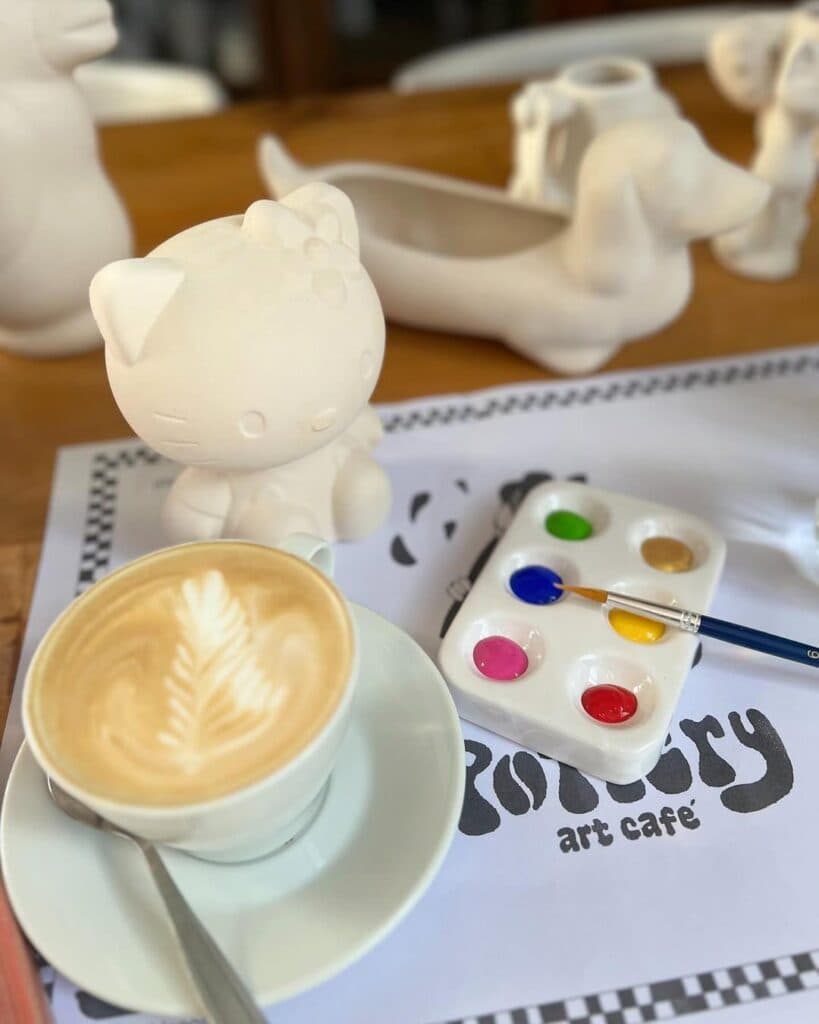 Café latte con arte, figura de Hello Kitty sin pintar, paleta de colores y logo Pottery Art Café sobre mesa de madera — imagen 1 de 4